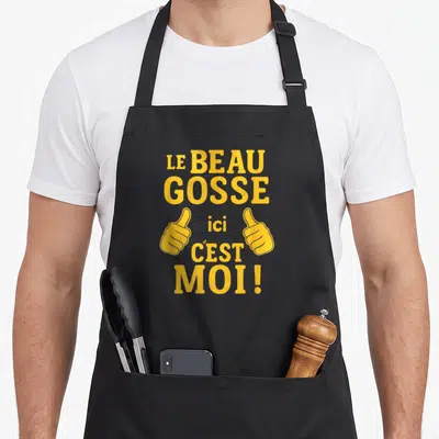 Tablier de cuisine noir avec texte jaune Le Beau Gosse Ici C'est Moi et deux mains faisant des pouces levés.