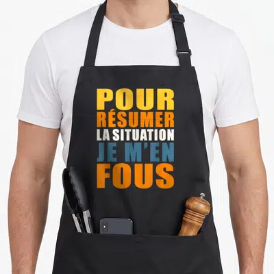 Tablier de cuisine noir avec l'inscription je m'en fous pour résumer la situation en lettres orange et bleues.