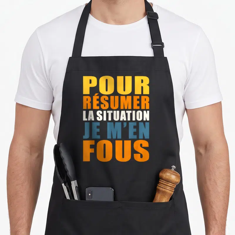 Tablier de cuisine noir avec l'inscription je m'en fous pour résumer la situation en lettres orange et bleues.