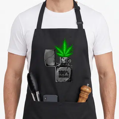 Tablier de cuisine noir avec illustration d'un briquet vintage crachant une feuille de cannabis verte et le texte 100% ORGANIC.