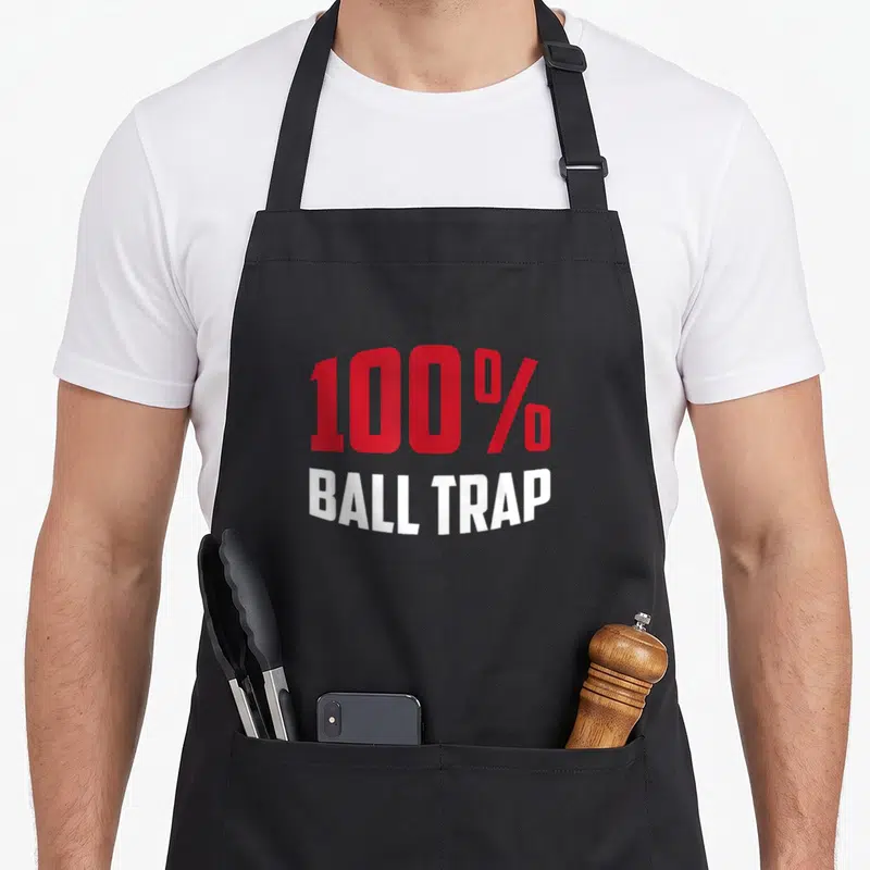 Tablier de cuisine noir avec l'inscription 100% Ball Trap en lettres rouges et blanches au centre du torse.