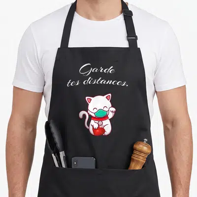 Tablier de cuisine noir avec illustration d'un chat blanc masqué vert tenant un cœur rouge sous le texte Garde tes distances.