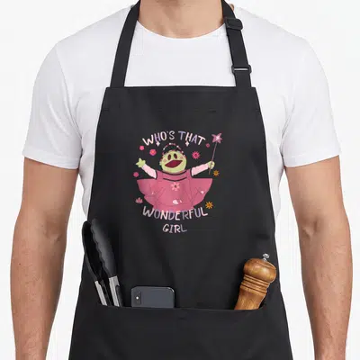 Tablier de cuisine noir avec illustration d'une marionnette rose en robe tenant une baguette magique entourée de fleurs et du texte Who's That Wonderful Girl.