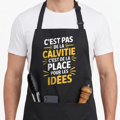 Tablier de cuisine noir avec inscription humoristique C'est pas de la calvitie c'est de la place pour les idées en lettres blanches et jaunes.