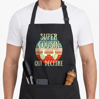 Tablier de cuisine noir avec inscription super cousin qui déchire et deux mains faisant un signe de pouce levé sur fond circulaire coloré.