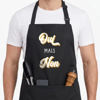 Tablier de cuisine noir avec l'inscription Oui Mais Non en lettres blanches et dorées stylisées.