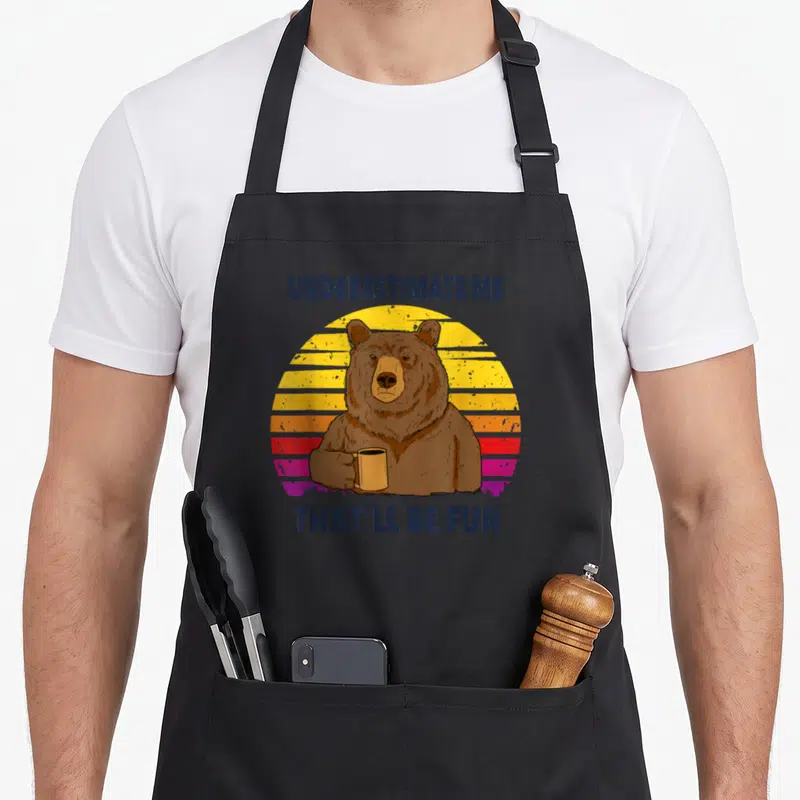 Tablier de cuisine noir avec illustration d'un ours tenant une tasse devant un soleil rétro multicolore.
