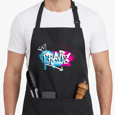 Tablier de cuisine noir avec le prénom Travis en graffiti blanc sur taches bleues et roses et une couronne.