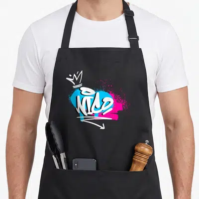 Tablier de cuisine noir avec le prénom Nico en style graffiti blanc sur fond bleu et rose vif, surmonté d'une couronne.