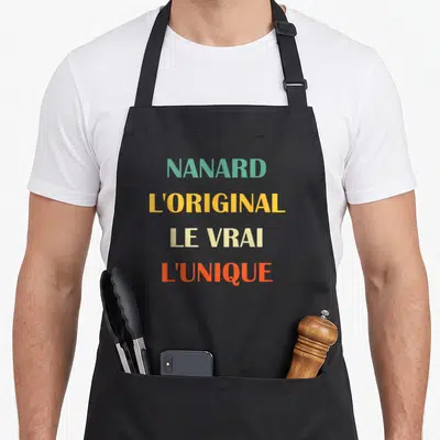 Tablier de cuisine noir avec les mots NANARD LORIGINAL LE VRAI LUNIQUE imprimés en couleurs vives sur le devant.