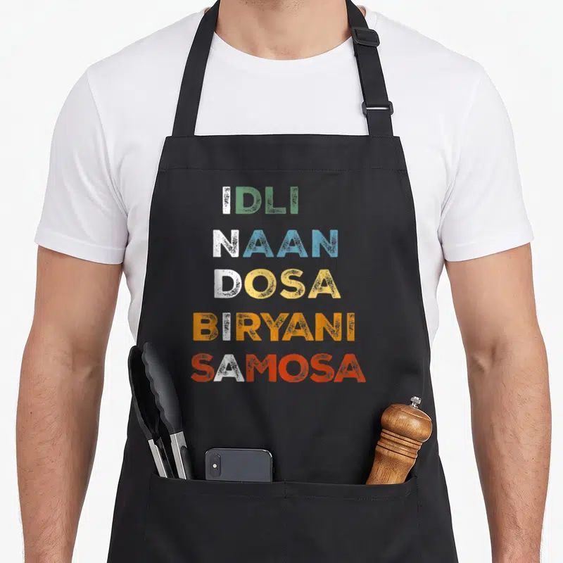 Tablier de cuisine noir avec les mots Idli Naan Dosa Biryani Samosa imprimés en lettres colorées vertes bleues jaunes oranges et rouges.