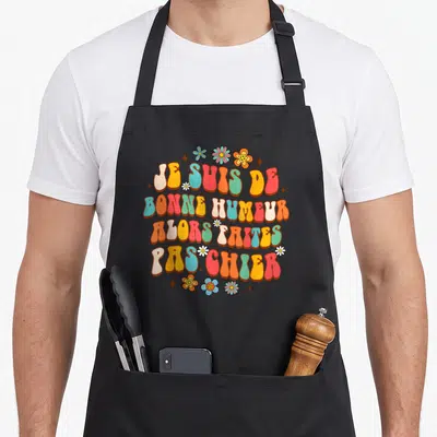 Tablier de cuisine noir avec inscription colorée Je Suis De Bonne Humeur Alors Faites Pas Chier entourée de fleurs stylisées.