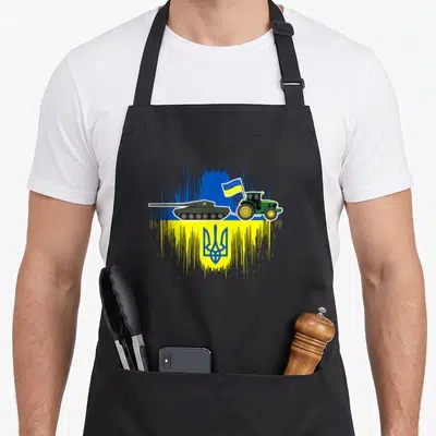 Tablier de cuisine noir avec illustration d'un tracteur vert tirant un char sur fond de drapeau ukrainien stylisé.