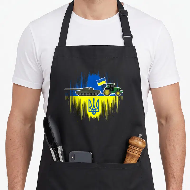 Tablier de cuisine noir avec illustration d'un tracteur vert tirant un char sur fond de drapeau ukrainien stylisé.