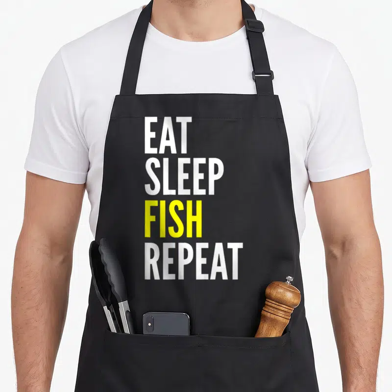 Tablier de cuisine noir avec l'inscription EAT SLEEP FISH REPEAT en lettres blanches et jaunes.