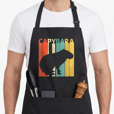Tablier de cuisine noir avec silhouette de capybara sur fond de crayons de couleur orange, beige, turquoise et jaune.
