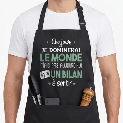 Tablier de cuisine noir avec l'inscription Un jour je dominerai le monde mais pas aujourd'hui il y a un bilan à sortir en vert et blanc.