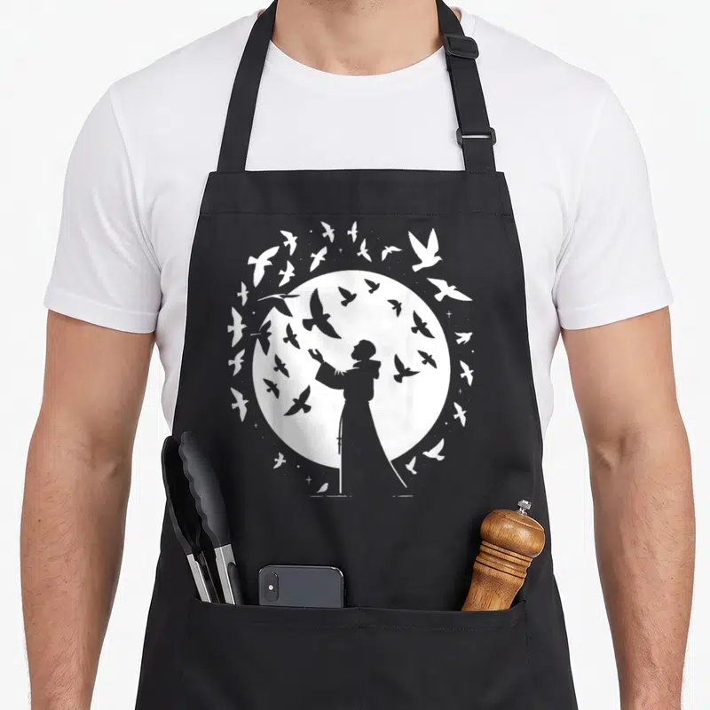 Tablier de cuisine noir avec silhouette blanche de Saint François d'Assise et oiseaux en vol autour d'un cercle lumineux.