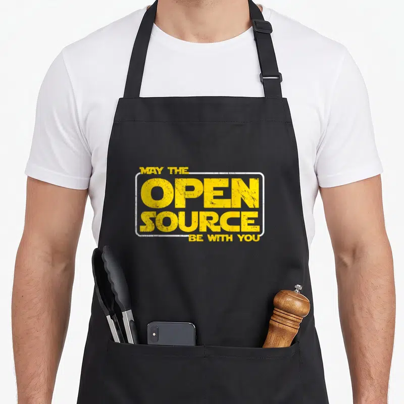 Tablier de cuisine noir avec l'inscription jaune May the Open Source Be with You dans un cadre blanc rectangulaire.