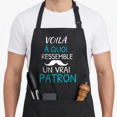 Tablier de cuisine noir avec le texte voilà à quoi ressemble un vrai patron et une moustache blanche stylisée.
