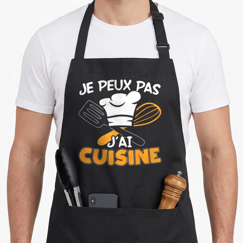 Tablier de cuisine noir avec toque, spatule et fouet orange portant l'inscription Je peux pas j'ai cuisine.