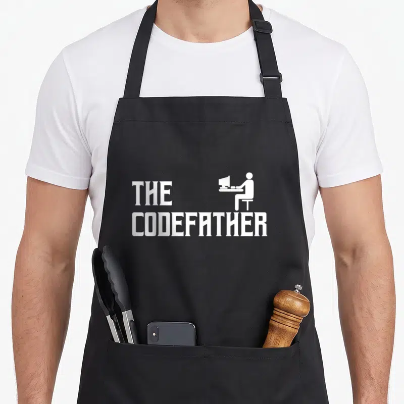 Tablier de cuisine noir avec le texte The Codefather et une silhouette blanche d'un homme devant un écran d'ordinateur.