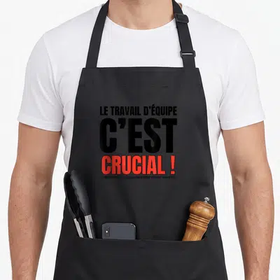 Tablier de cuisine noir avec le texte Le travail d'équipe c'est crucial en lettres noires et rouges.