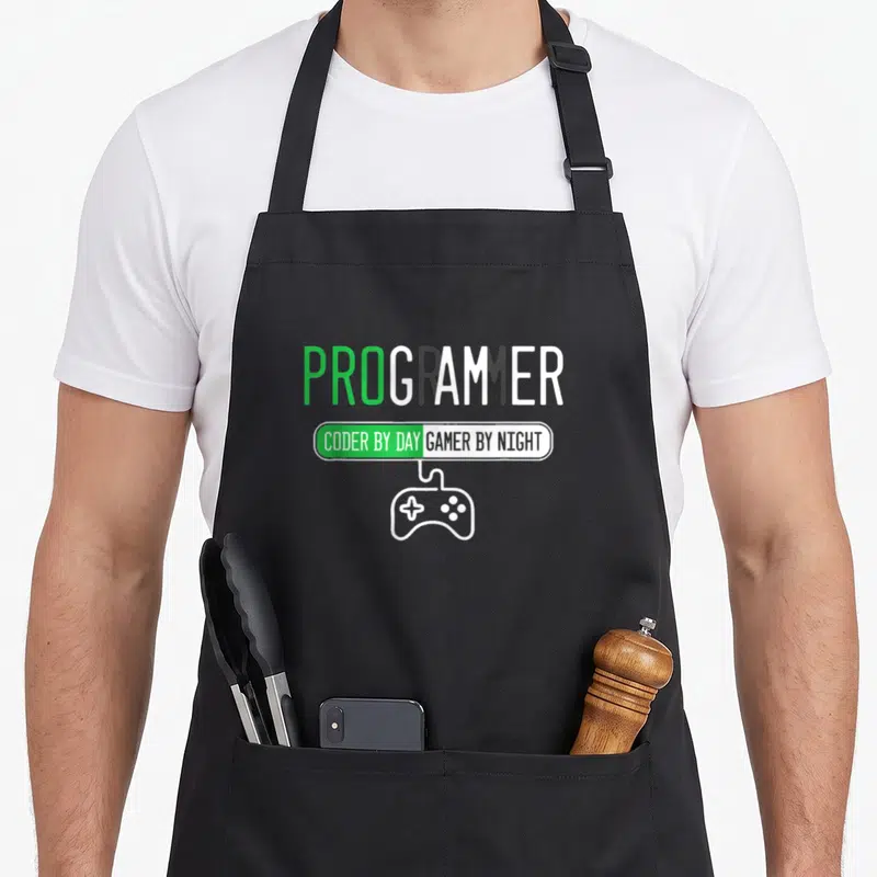Tablier de cuisine noir avec texte PROGRAMMER et icône manette de jeu vidéo verte et blanche au centre.