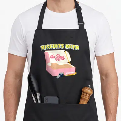 Tablier de cuisine noir avec illustration d'une boîte de biscuits rose, d'un sifflet et du texte Biscuits With The Boss en lettres jaunes.
