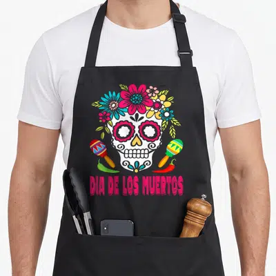 Tablier de cuisine noir avec crâne Dia de los Muertos entouré de fleurs, maracas et piments en bas.
