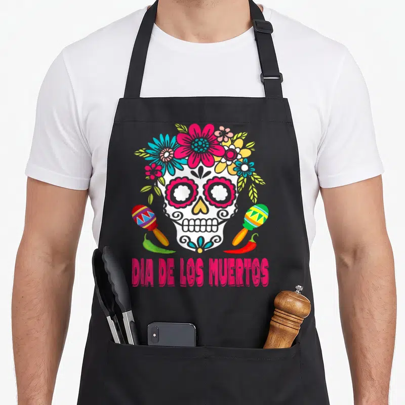 Tablier de cuisine noir avec crâne Dia de los Muertos entouré de fleurs, maracas et piments en bas.