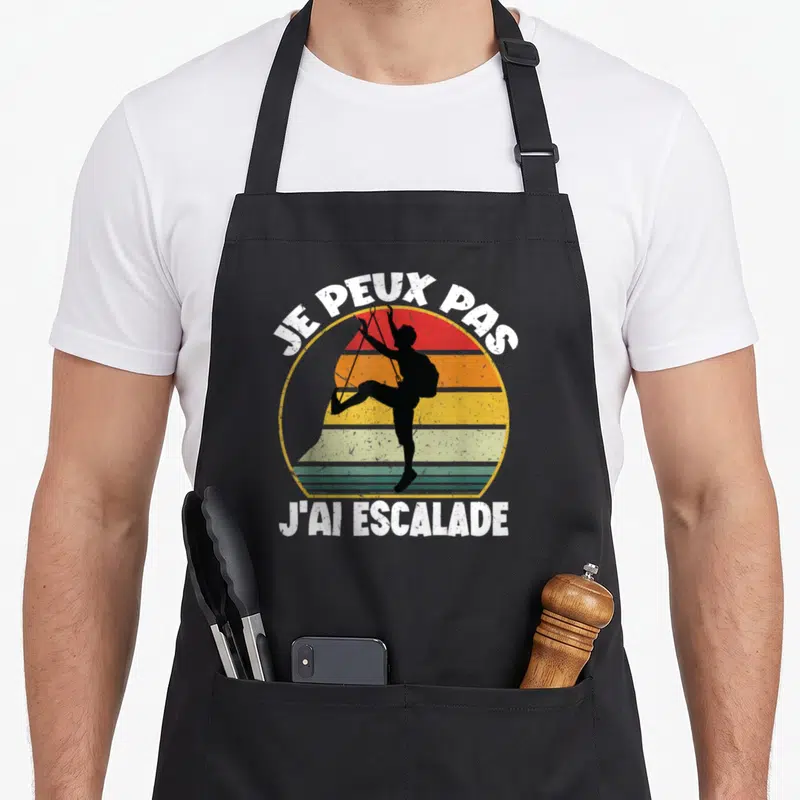 Tablier de cuisine noir avec silhouette grimpeur sur fond de soleil couchant multicolore et texte blanc.