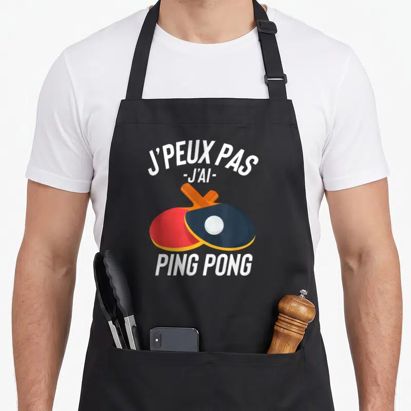 Tablier de cuisine noir avec deux raquettes de ping-pong rouge et noire croisées et une balle blanche au centre.