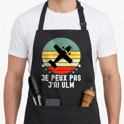 Tablier de cuisine noir avec silhouette d'avion ULM et texte blanc sur fond de soleil couchant coloré.