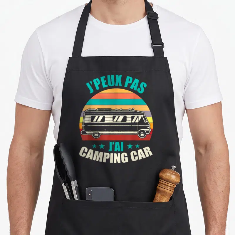 Tablier de cuisine noir avec inscription jpeux pas jai camping car et illustration de van colorée sur fond de soleil dégradé.