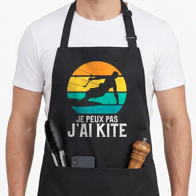 Tablier de cuisine noir avec silhouette de kitesurfeur sur fond de soleil coloré et texte blanc.