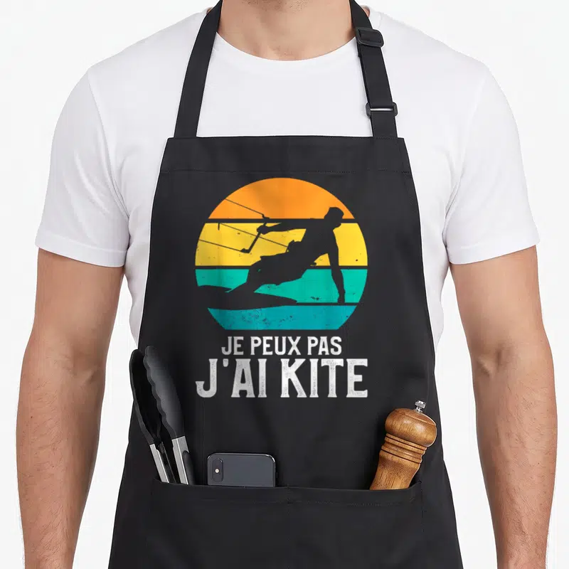 Tablier de cuisine noir avec silhouette de kitesurfeur sur fond de soleil coloré et texte blanc.