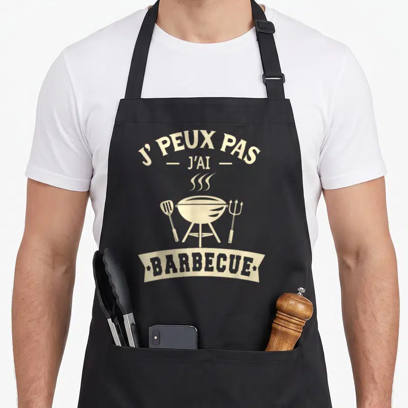 Tablier de cuisine noir avec illustration vintage d'un barbecue et ustensiles de cuisson beige et gris.