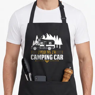 Tablier de cuisine noir avec illustration d'une caravane blanche, des sapins et le texte J'peux pas j'ai camping car en blanc et orange.
