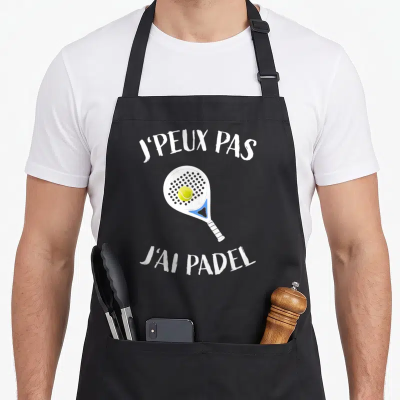 Tablier de cuisine noir avec une raquette de padel, une balle jaune et le texte j'peux pas j'ai padel imprimé en blanc.