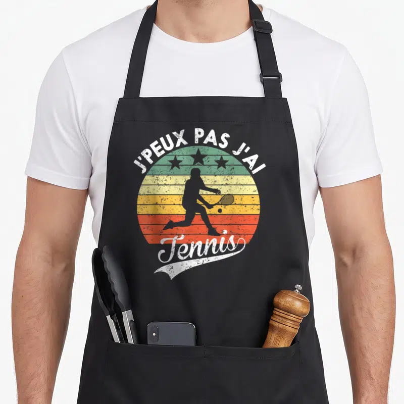 Tablier de cuisine noir avec silhouette de joueur de tennis et texte J'peux pas j'ai tennis sur fond de soleil vintage.