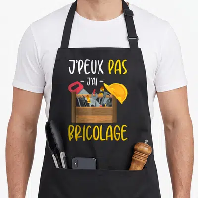 Tablier de cuisine noir avec illustration boîte d'outils et texte J'peux pas j'ai bricolage en lettres jaunes et blanches.