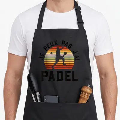 Tablier de cuisine noir avec silhouette de joueur de padel devant un soleil rayé orange et jaune, texte en arc de cercle.