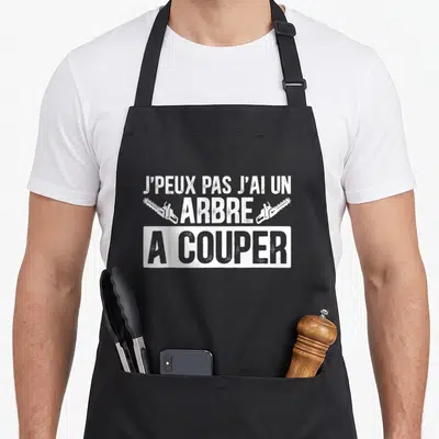 Tablier de cuisine noir avec inscription j'peux pas j'ai un arbre à couper et deux tronçonneuses blanches.