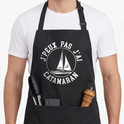 Tablier de cuisine noir avec logo circulaire blanc représentant un catamaran et le texte j'peux pas j'ai catamaran.