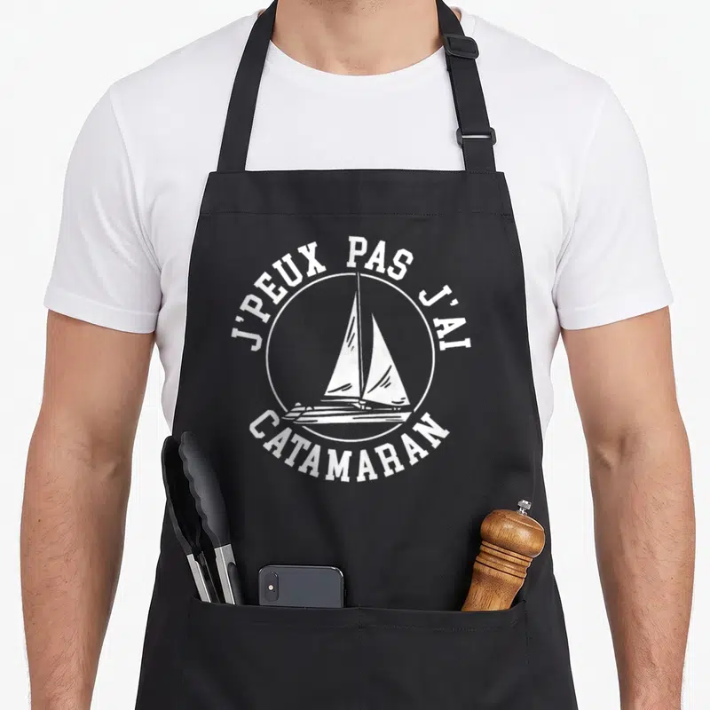 Tablier de cuisine noir avec logo circulaire blanc représentant un catamaran et le texte j'peux pas j'ai catamaran.