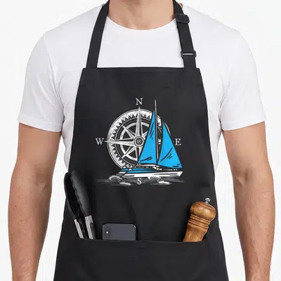 Tablier de cuisine noir avec catamaran aux voiles bleues devant une boussole blanche.