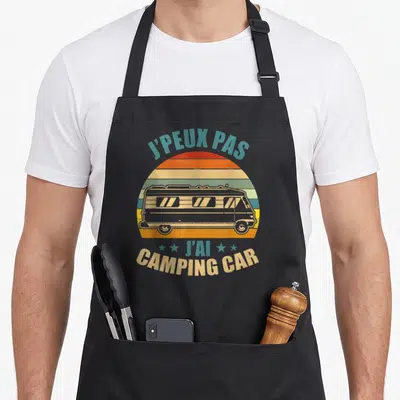 Tablier de cuisine noir avec illustration d'un camping-car vintage devant un soleil multicolore et le texte j'peux pas j'ai camping car.
