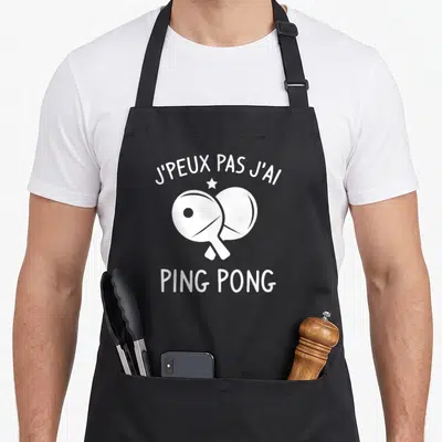 Tablier de cuisine noir avec deux paddles de tennis de table blancs croisés et le texte j'peux pas j'ai ping pong.