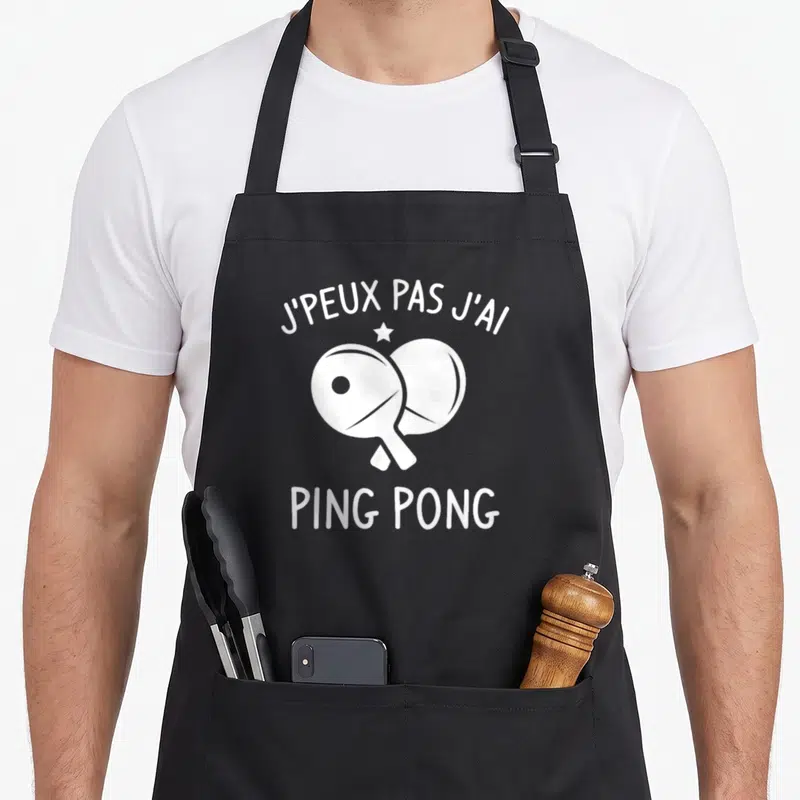 Tablier de cuisine noir avec deux paddles de tennis de table blancs croisés et le texte j'peux pas j'ai ping pong.